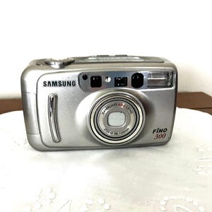 Könnte beinhalten: Silberne Samsung Fino 300 Kamera. Die Kamera hat ein Zoomobjektiv und einen eingebauten Blitz. Die Kamera ist rechteckig mit abgerundeten Kanten und einer silbernen Oberfläche. Das Samsung-Logo ist oben links zu sehen.