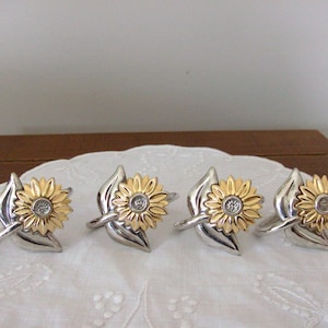 Könnte beinhalten: Set aus vier Serviettenringen in Form von Sonnenblumen in Silber und Gold. Jeder Ring zeigt eine stilisierte Sonnenblume mit einem klaren Strassstein in der Mitte.