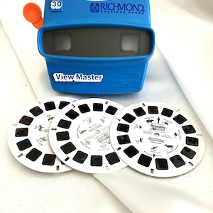 Puede incluir: Un visor View-Master 3D azul con una palanca naranja y tres discos blancos. El visor tiene el texto "View-Master" y "Richmond American Homes". Los discos tienen pequeñas imágenes y texto.