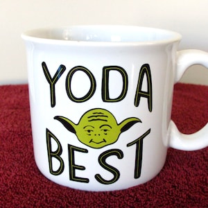 K&ouml;nnte beinhalten: Wei&szlig;e Keramik-Tasse mit einem gr&uuml;nen Yoda-Motiv und dem Schriftzug "YODA BEST" in Schwarz. Die Tasse hat einen gebogenen Henkel. Ein lustiges Geschenk f&uuml;r Star Wars-Fans, geeignet f&uuml;r Tee, Kaffee oder andere Hei&szlig;getr&auml;nke.