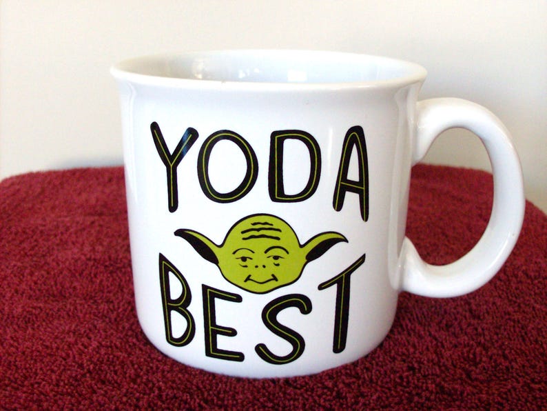 K&ouml;nnte beinhalten: Wei&szlig;e Keramik-Tasse mit einem gr&uuml;nen Yoda-Motiv und dem Schriftzug "YODA BEST" in Schwarz. Die Tasse hat einen gebogenen Henkel. Ein lustiges Geschenk f&uuml;r Star Wars-Fans, geeignet f&uuml;r Tee, Kaffee oder andere Hei&szlig;getr&auml;nke.