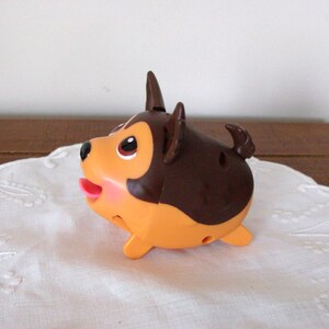 May include: A small, rounded plastic toy dog with a dark brown back and head, and a light orange face, chest, and underbelly. The figure features large brown eyes, a black nose, a small pink tongue, and pointed ears, with a small tail.