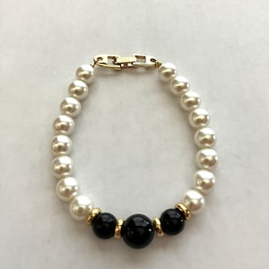 May include: A pearl bracelet with a gold clasp. The bracelet features alternating white and black beads. The black beads are larger than the white beads and are accented with gold-colored spacers.