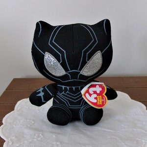 May include: A black plush toy resembling the Black Panther superhero. The toy has silver glitter eyes and light blue accents. It is sitting on a white, scalloped doily. A red and white tag is attached.