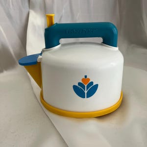 May include: A white plastic toy kettle with a blue handle and spout, and a yellow base and spout. The kettle has a blue and orange floral design on the front and the Fisher-Price logo on the handle.