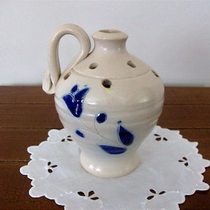 Op de afbeelding: Een crèmekleurige keramische kruik met een handvat en een blauw bloemmotief. De kruik heeft verschillende gaten aan de bovenkant en staat op een witte, geschulpte doily. De kruik staat op een donkerbruin houten oppervlak.