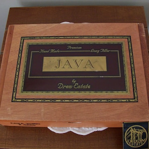 Peut inclure: Une boîte en bois avec une étiquette dorée qui indique "Premium Hand Made Long Filler JAVA by Drew Estate".