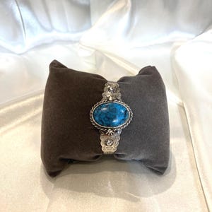 Puede incluir: Brazalete plateado con una piedra turquesa ovalada. La piedra está engastada en un marco plateado detallado con un borde en forma de cuerda. El brazalete tiene elementos decorativos en la banda y se muestra sobre un cojín de terciopelo gris oscuro.