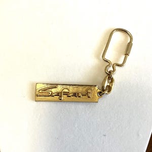 May include: A gold-colored metal keychain with the word "Safari" engraved on a rectangular plate. The keychain has a simple design with a key ring attached to the plate by a small chain.
