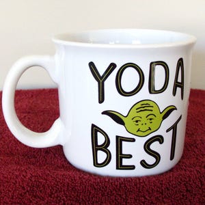 K&ouml;nnte beinhalten: Wei&szlig;er Keramikbecher mit gebogenem Henkel. Der Becher zeigt die Worte "YODA BEST" in schwarzen Buchstaben mit einer gr&uuml;nen Yoda-Charakter-Illustration. Der Becher steht auf einer roten Oberfl&auml;che.