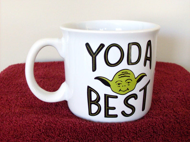 K&ouml;nnte beinhalten: Wei&szlig;er Keramikbecher mit gebogenem Henkel. Der Becher zeigt die Worte "YODA BEST" in schwarzen Buchstaben mit einer gr&uuml;nen Yoda-Charakter-Illustration. Der Becher steht auf einer roten Oberfl&auml;che.