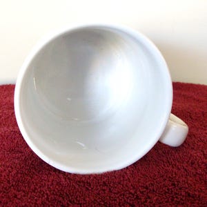 K&ouml;nnte beinhalten: Eine wei&szlig;e Keramik-Tasse mit rundem Innenraum und Henkel. Die Tasse steht vor einem roten, strukturierten Hintergrund. Die Tasse ist leer und sauber.