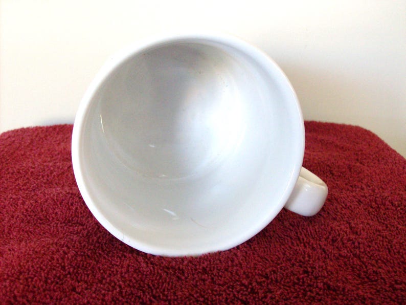 K&ouml;nnte beinhalten: Eine wei&szlig;e Keramik-Tasse mit rundem Innenraum und Henkel. Die Tasse steht vor einem roten, strukturierten Hintergrund. Die Tasse ist leer und sauber.