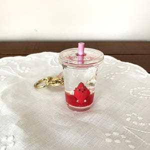 Puede incluir: Un llavero de taza de bebida transparente en miniatura con un dije rojo en forma de fresa en el interior. La taza tiene una pajita rosa y un accesorio de llavero dorado. La bebida parece estar sobre un paño blanco bordado.