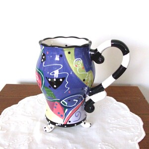 May include: A colorful ceramic mug with a whimsical design. The mug features a blue body with abstract shapes in pink, green, and white. The handle is black and white striped. The mug has a scalloped rim and sits on three small feet.