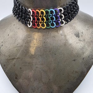 May include: Black chainmail choker necklace with a rainbow accent. The choker features a series of black metal rings, with a row of colorful rings in white, red, orange, yellow, green, blue, and purple. The necklace is displayed on a metal bust.