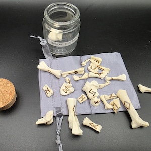 May include: A clear glass jar with a label reading "Beaver Toe Bones" next to a cork stopper and a gray pouch containing bone-shaped objects with etched markings. The objects are arranged on a dark surface.