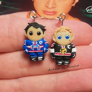 May include: Handmade polymer clay earrings featuring two miniature hockey player figurines. One player wears a blue jersey, the other a black jersey. Both have detailed facial features and are suspended from silver-tone earring hooks. The text "Artifex de Mundus" is visible.
