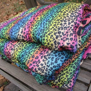 May include: A folded blanket with a vibrant rainbow leopard print. The blanket features a spectrum of colors, including pink, yellow, blue, and green, with black spots. It is resting on a wooden bench, showcasing its bold design.