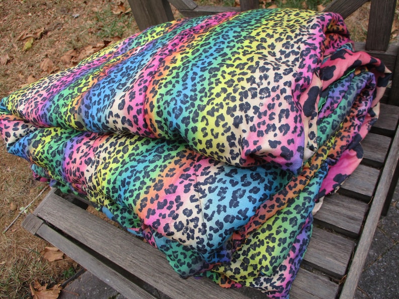 May include: A folded blanket with a vibrant rainbow leopard print. The blanket features a spectrum of colors, including pink, yellow, blue, and green, with black spots. It is resting on a wooden bench, showcasing its bold design.