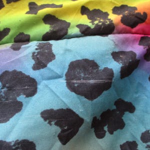 May include: Close-up of a fabric with a vibrant rainbow gradient background. Large black spots are scattered across the fabric. The colors transition from yellow to green, blue, purple, and pink.