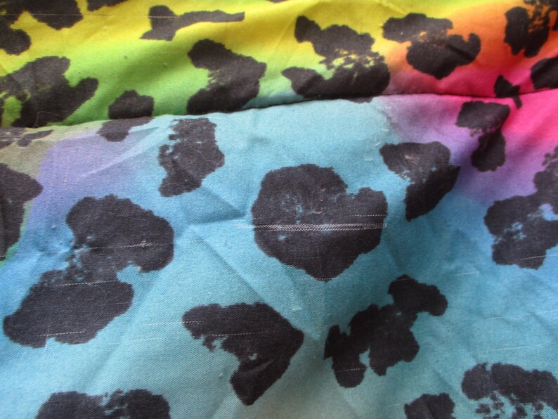 May include: Close-up of a fabric with a vibrant rainbow gradient background. Large black spots are scattered across the fabric. The colors transition from yellow to green, blue, purple, and pink.