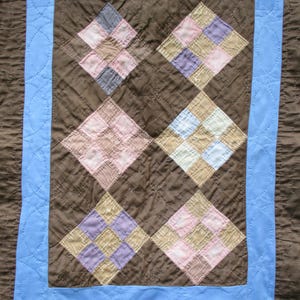 Op de afbeelding: Een handgemaakte quilt met een bruin midden en een lichtblauwe rand. Het midden heeft zes ruitvormige patches in verschillende pastelkleuren, waaronder roze, paars en beige. De quilt heeft een gewatteerd patroon.