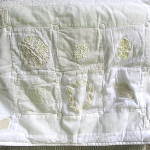 May include: A white rectangular quilted pillow sham featuring a grid pattern of stitched squares. Each square contains unique off-white embroidered motifs, including floral patterns, geometric shapes, and a central laurel wreath design.