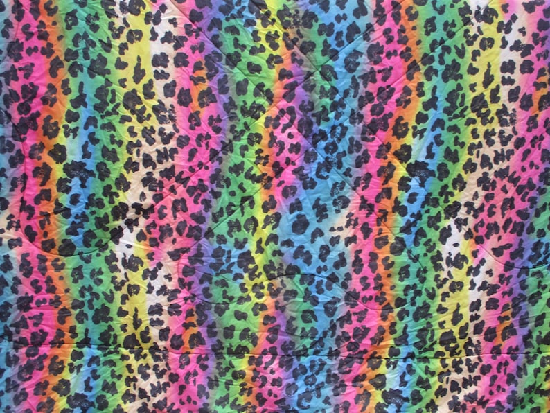 May include: A vibrant textile featuring a rainbow-colored background with vertical stripes. The design is overlaid with a pattern of black leopard spots, creating a bold and colorful animal print. The fabric has a slightly wrinkled texture.