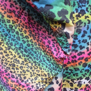 May include: Close-up of a fabric with a vibrant rainbow leopard print. The design features a gradient of colors, including pink, yellow, green, and blue, with black leopard spots. The fabric is folded, showing the texture and pattern.