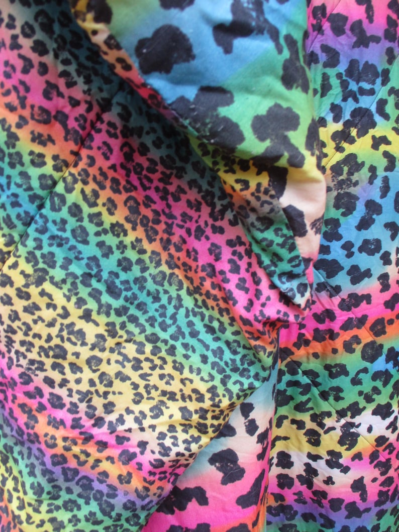 May include: Close-up of a fabric with a vibrant rainbow leopard print. The design features a gradient of colors, including pink, yellow, green, and blue, with black leopard spots. The fabric is folded, showing the texture and pattern.