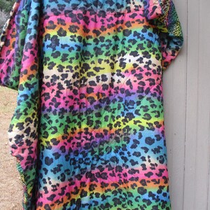 May include: A vibrant, rainbow-colored blanket with a leopard print design. The blanket features horizontal stripes of various colors, including pink, orange, yellow, green, and blue, with black leopard spots. The blanket is draped over a wooden surface.