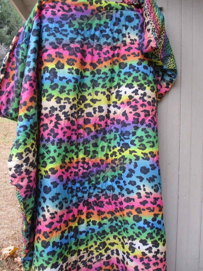 May include: A vibrant, rainbow-colored blanket with a leopard print design. The blanket features horizontal stripes of various colors, including pink, orange, yellow, green, and blue, with black leopard spots. The blanket is draped over a wooden surface.