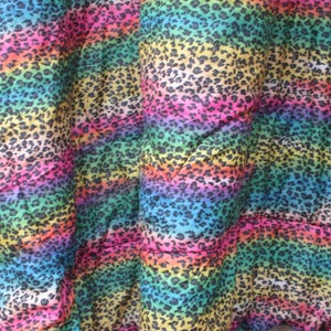 May include: A close-up of a fabric with a rainbow leopard print pattern. The colors transition from blue to pink, yellow, and green, with black spots. The fabric is folded, showing the texture and design.