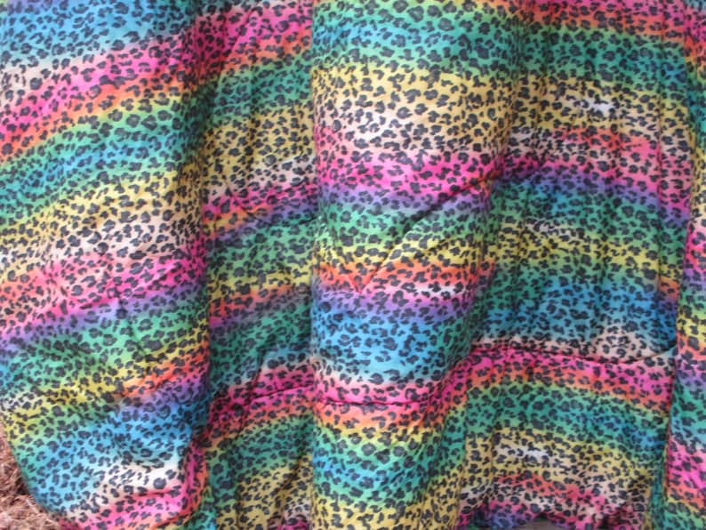 May include: A close-up of a fabric with a rainbow leopard print pattern. The colors transition from blue to pink, yellow, and green, with black spots. The fabric is folded, showing the texture and design.