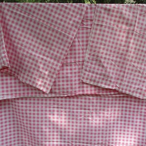 May include: A large piece of fabric featuring a classic pink and white gingham check pattern. The textile displays a consistent grid of small squares, with the light pink and white colors alternating throughout the entire surface of the material.