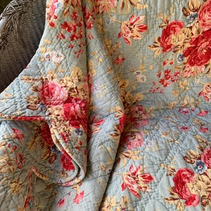 May include: A quilted blanket featuring a light blue background with a repeating floral pattern of red and pink roses, along with smaller cream and tan blossoms and green leaves. The fabric has a visible diamond-shaped stitched texture throughout.