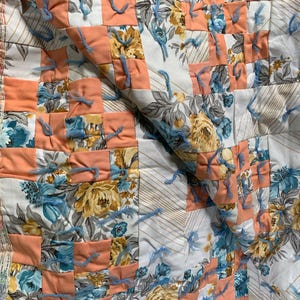 Puede incluir: Un edredón hecho a mano con un diseño de patchwork. El edredón presenta cuadrados de tela color melocotón y estampados florales en tonos de azul, amarillo y blanco. Se utiliza hilo azul para conectar los cuadrados, agregando textura y detalle al textil.