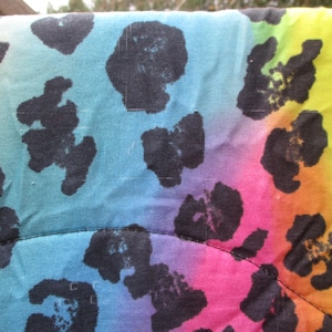 May include: Close-up of a fabric with a rainbow gradient background and black leopard print spots. The colors transition from blue to pink, orange, and green. The fabric has a quilted appearance.