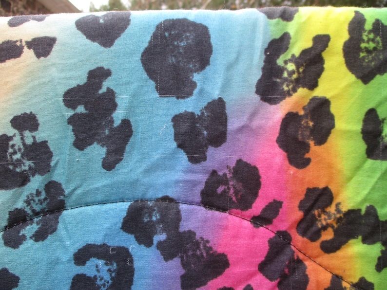 May include: Close-up of a fabric with a rainbow gradient background and black leopard print spots. The colors transition from blue to pink, orange, and green. The fabric has a quilted appearance.
