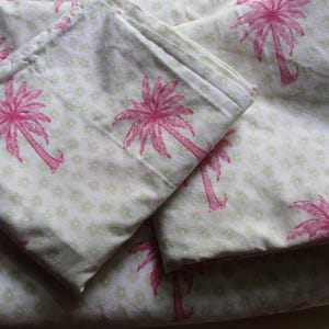 May include: A folded fabric sheet featuring a repeating pattern of bright pink palm trees set against a light cream background. The fabric is also decorated with a subtle, small-scale light green floral print dispersed between the palm trees.