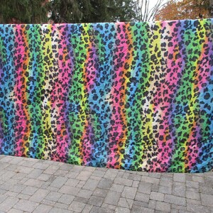 May include: A vibrant, colorful textile with a rainbow leopard print pattern. The fabric features vertical stripes of pink, orange, yellow, green, blue, and purple, with black leopard spots. The textile is hanging outdoors.