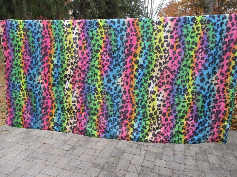 May include: A vibrant, colorful textile with a rainbow leopard print pattern. The fabric features vertical stripes of pink, orange, yellow, green, blue, and purple, with black leopard spots. The textile is hanging outdoors.