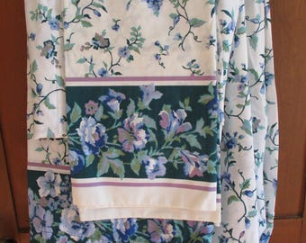 May include: White floral bed sheets with a dark green border featuring blue and purple flowers. The sheets have a repeating pattern of blue flowers and green vines on a white background. The sheets are folded and hanging.