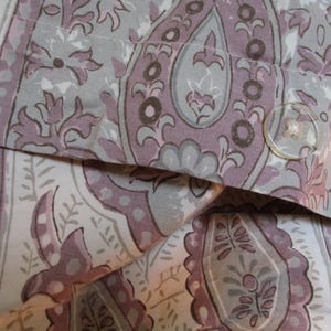 Peut inclure: Gros plan sur un tissu à motifs avec un motif paisley dans les tons de violet, gris et blanc. Le tissu est plié sur lui-même, créant un effet de superposition. Un petit bouton transparent est visible sur la couche supérieure du tissu.