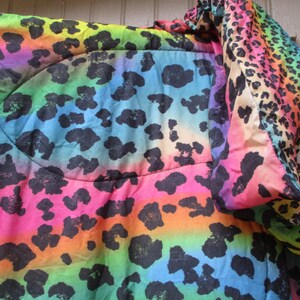 May include: A vibrant, rainbow-colored blanket with a leopard print design. The blanket features a gradient of colors, including pink, orange, yellow, green, blue, and purple, with black leopard spots. The fabric appears soft and quilted.