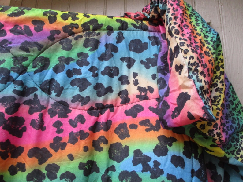May include: A vibrant, rainbow-colored blanket with a leopard print design. The blanket features a gradient of colors, including pink, orange, yellow, green, blue, and purple, with black leopard spots. The fabric appears soft and quilted.