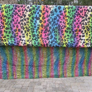 May include: Two colorful leopard print fabrics. The top fabric has a rainbow gradient background with black spots. The bottom fabric has vertical rainbow stripes with black spots. The fabrics are displayed outdoors.