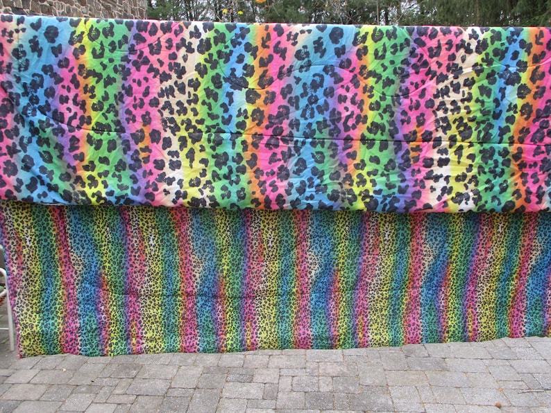 May include: Two colorful leopard print fabrics. The top fabric has a rainbow gradient background with black spots. The bottom fabric has vertical rainbow stripes with black spots. The fabrics are displayed outdoors.