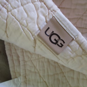 May include: Close-up of a cream-colored quilted blanket. The blanket has a diamond pattern and a soft texture. A rectangular leather tag with the brand name "UGG" in black lettering is sewn on the corner of the blanket.